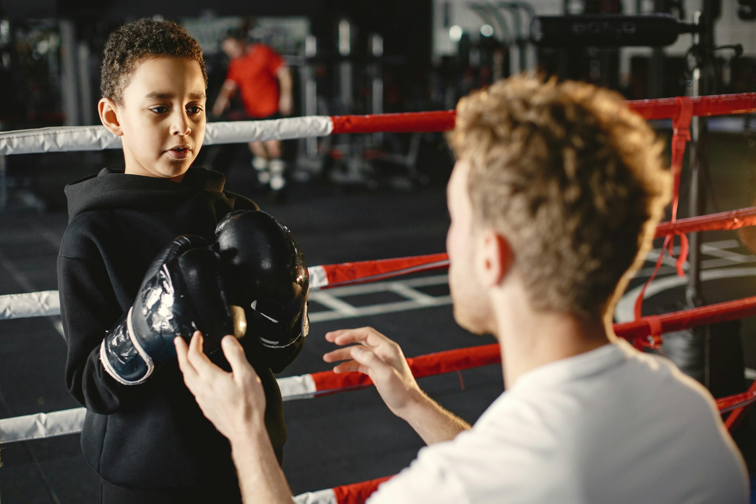 A child in boxing gloves trains with a coach in a gym boxing ring, focused on learning skills.