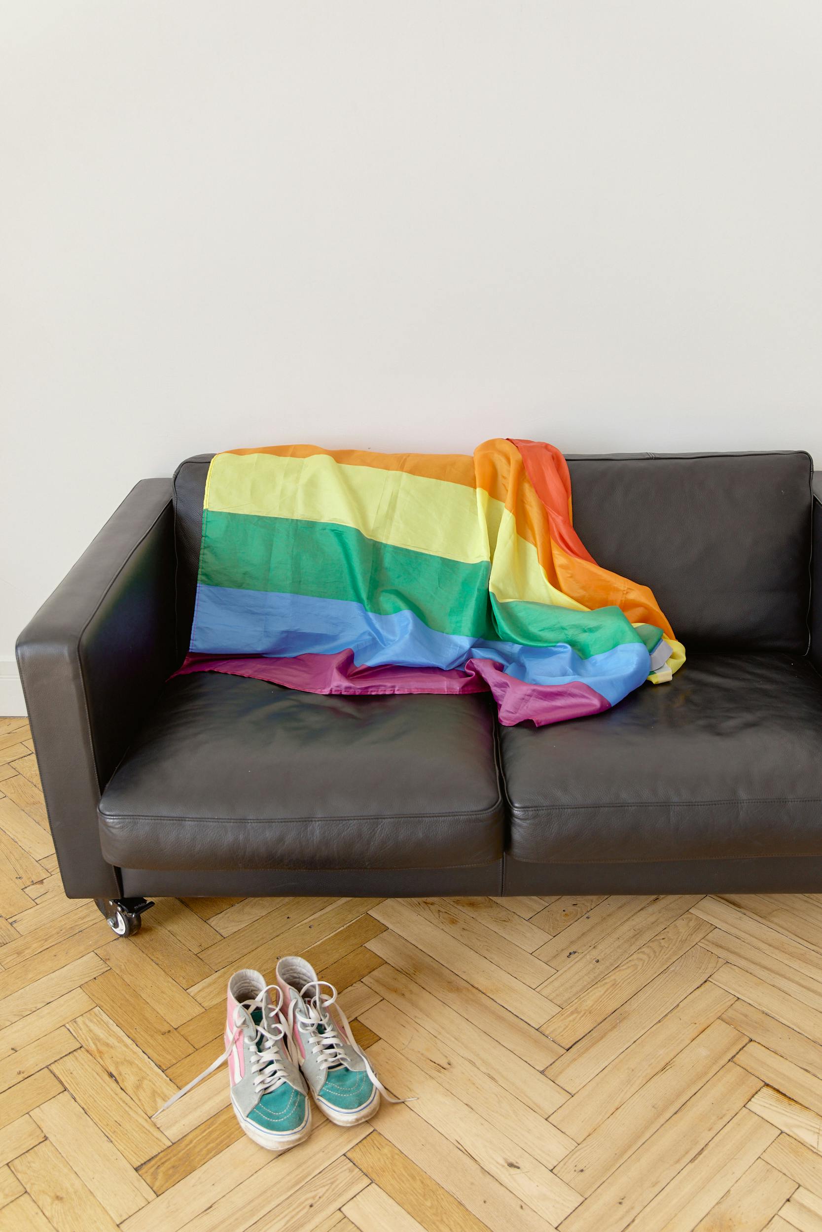 Rainbow pride flag draped on a leather sofa with sneakers on wooden floor.